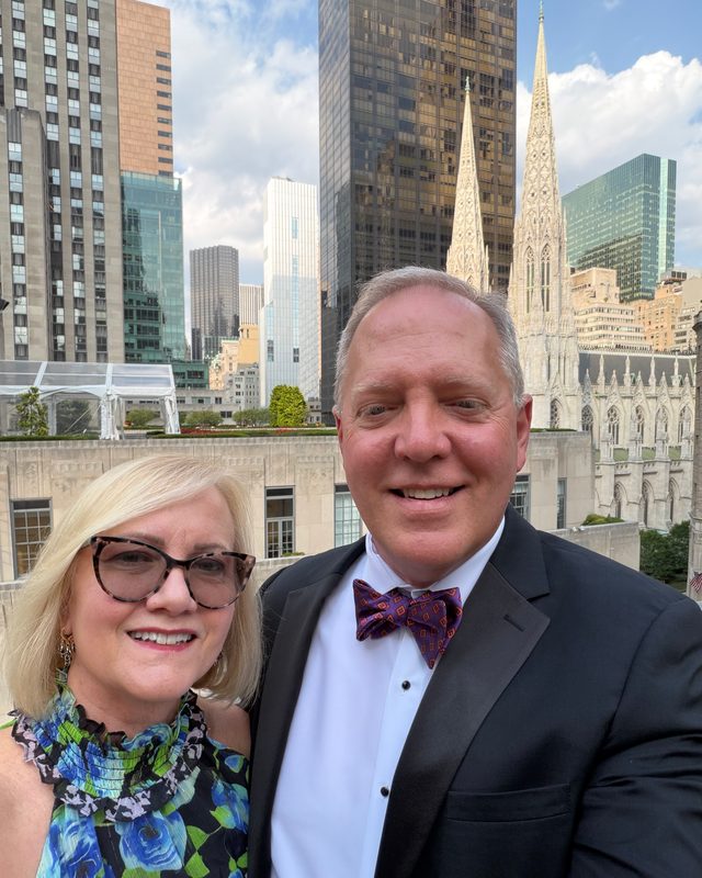 Scott Allison in a tuxedo beside a woman in a floral dress, with skyscrapers and a cathedral behind them
