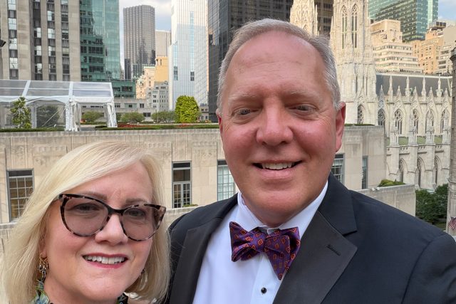 Scott Allison in a tuxedo beside a woman in a floral dress, with skyscrapers and a cathedral behind them