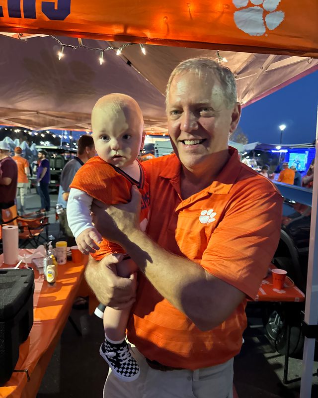 Scott Allison holding a baby under an orange tailgate tent